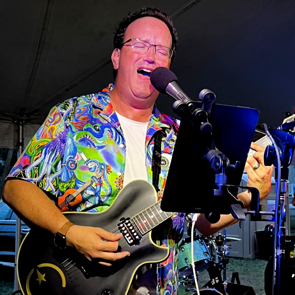 Person playing guitar and singing into a microphone with a colorful shirt.