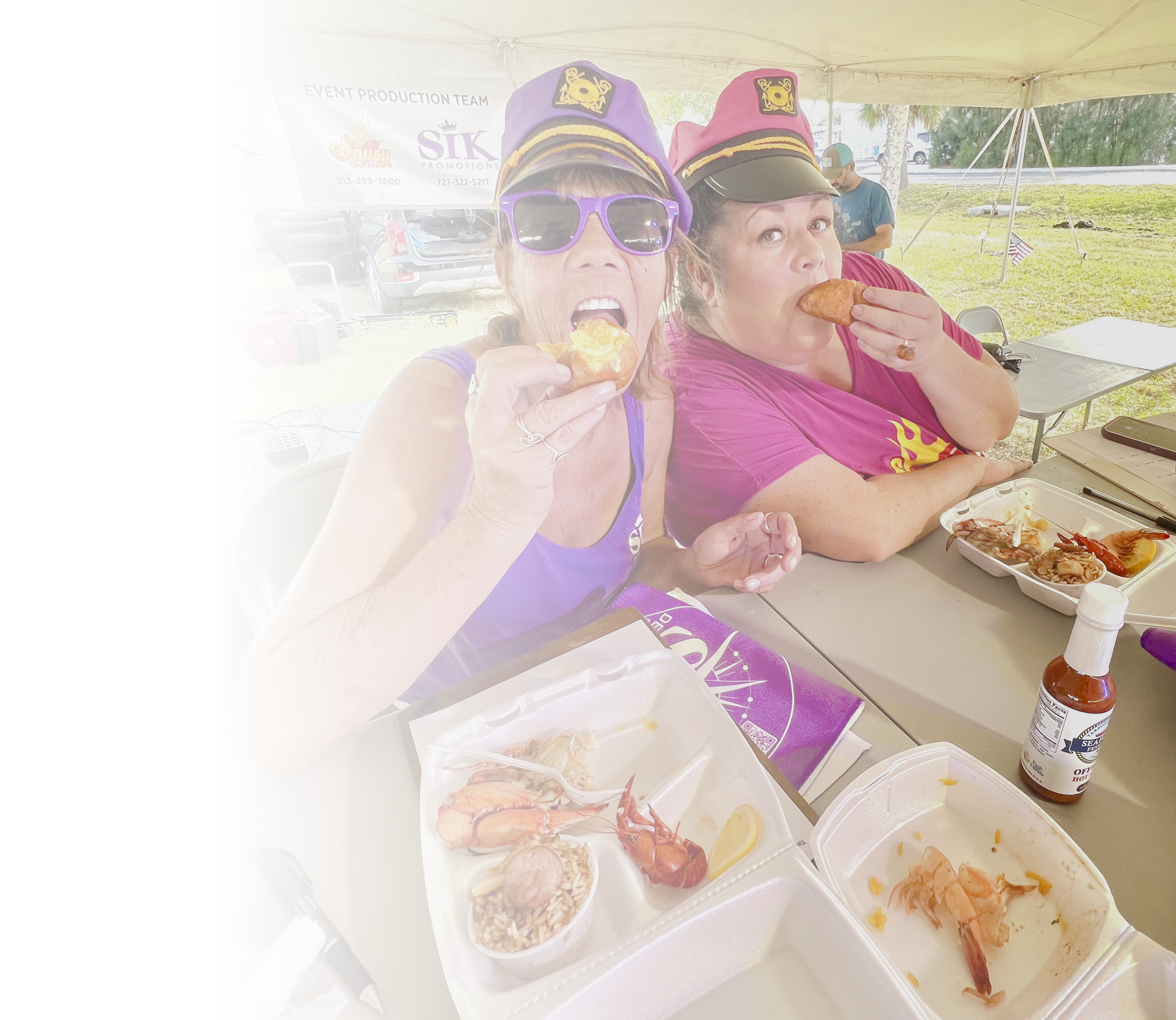 Two people eating at a food stand with food containers and a bottle of hot sauce.